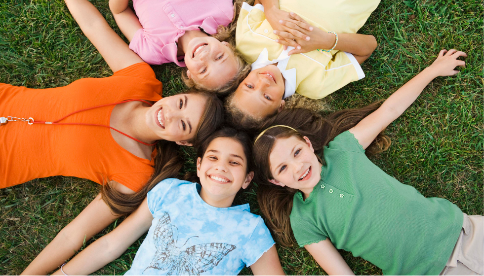 Top down image of five girls laying in grass, heads touching, all smiling