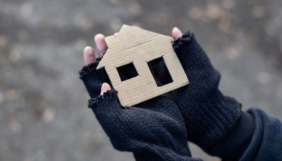 Dark image of hands in tattered gloves holding small piece of carboard in shape of a house
